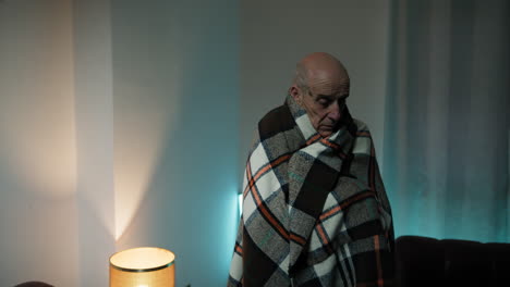 elderly man wraps himself in a blanket at home due to the cold