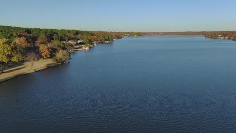 Vista-Aérea-A-Baja-Altitud-De-Las-Casas-A-Lo-Largo-De-La-Orilla-Del-Lago-Con-Un-Telón-De-Fondo-De-Colores-Otoñales