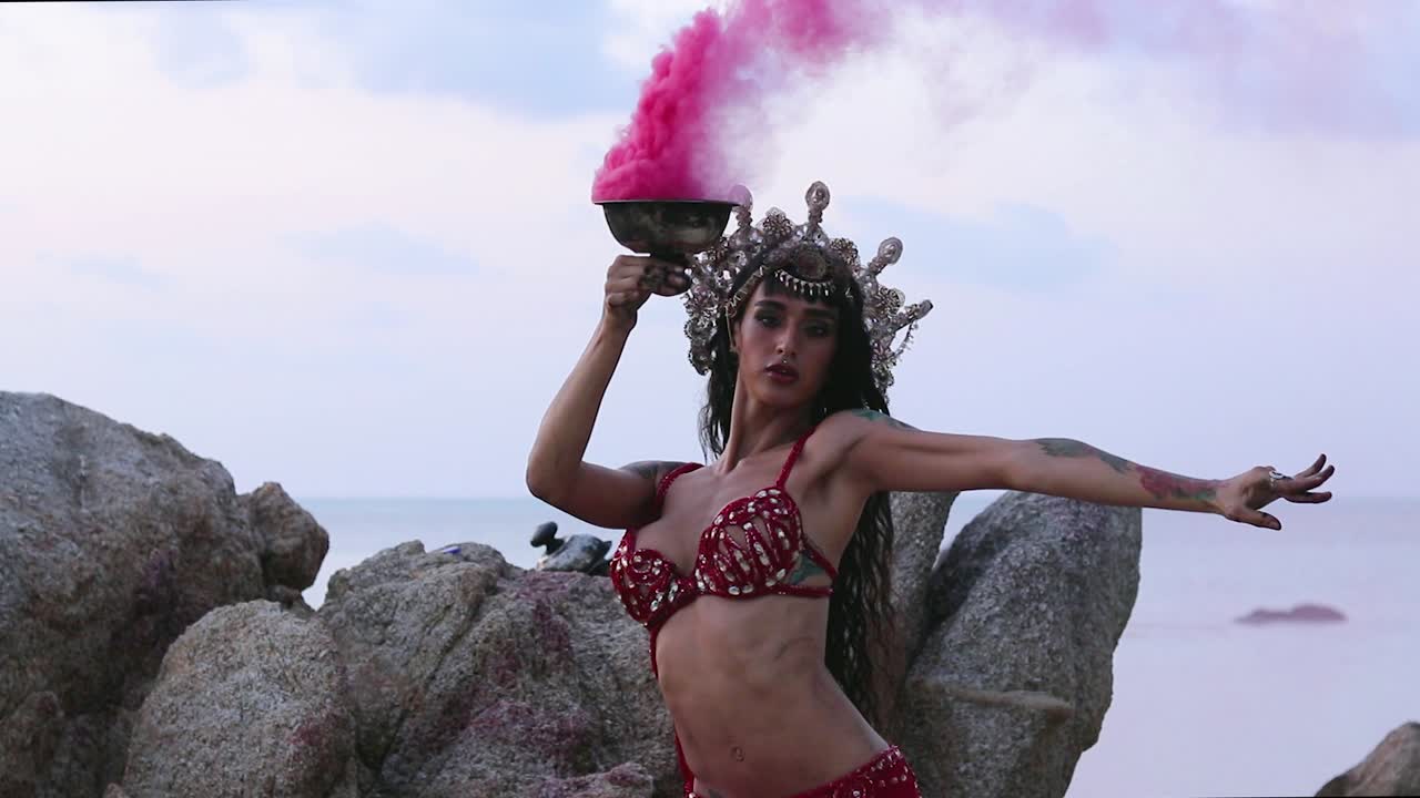 Beautiful, young and sexy belly dancer performing rhythmic moves with pink color smoke bomb, on the beach. Slow motion