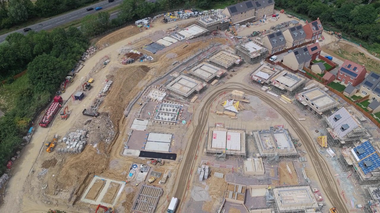 Nuove case in costruzione mostrano fondamenta e strutture in evoluzione in una tranquilla zona suburbana durante il giorno, con macchinari pesanti visibili sul terreno polveroso vicino a una strada principale
