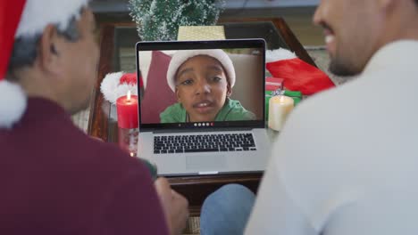 padre y hijo biraciales sonrientes usando una computadora portátil para una videollamada de navidad con el niño en la pantalla
