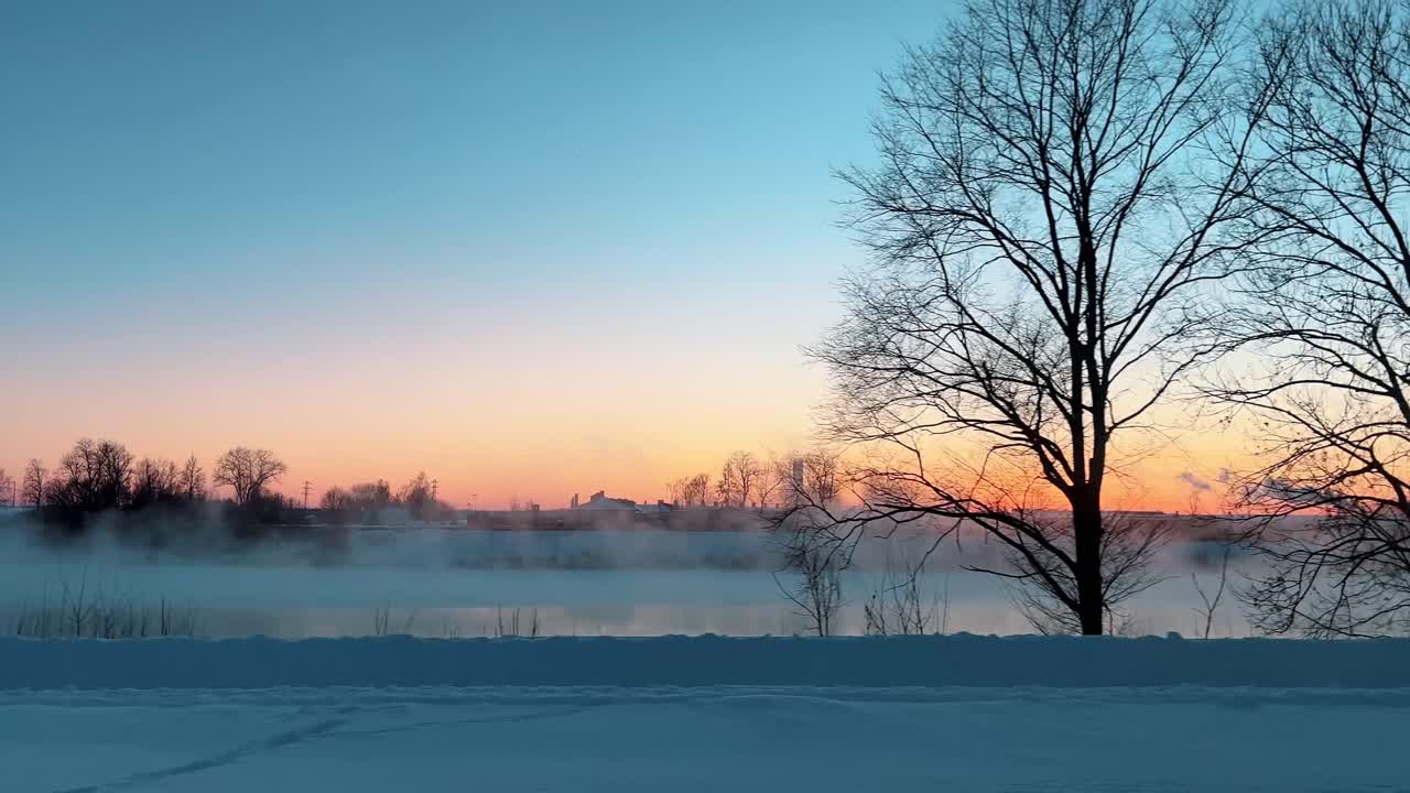 city park covered in snow during a cold winter sunrise, with thick fog rising from the river
