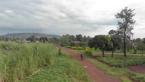 Aerial-shot-tracking-from-behind-a-African-man-wearing-a-helmet-on-a-motorcycle-riding-on-a-dirt-road-through-a-rural-village