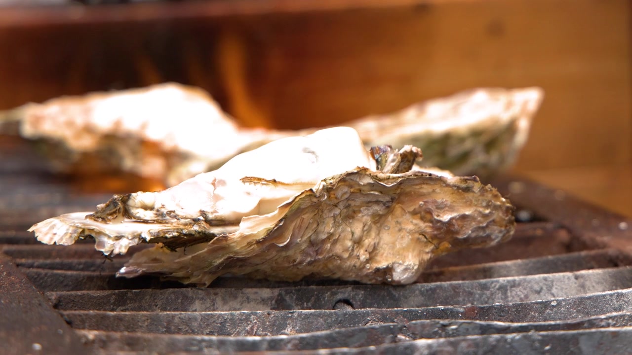 Close-up shot of large oysters cooking on a metal grill with flickering orange flames.