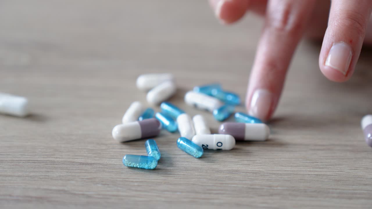 Close-up shot of a hand slowly arranging a pile of blue and white capsules on a wooden table, evoking themes of health, medication, and personal care decisions.