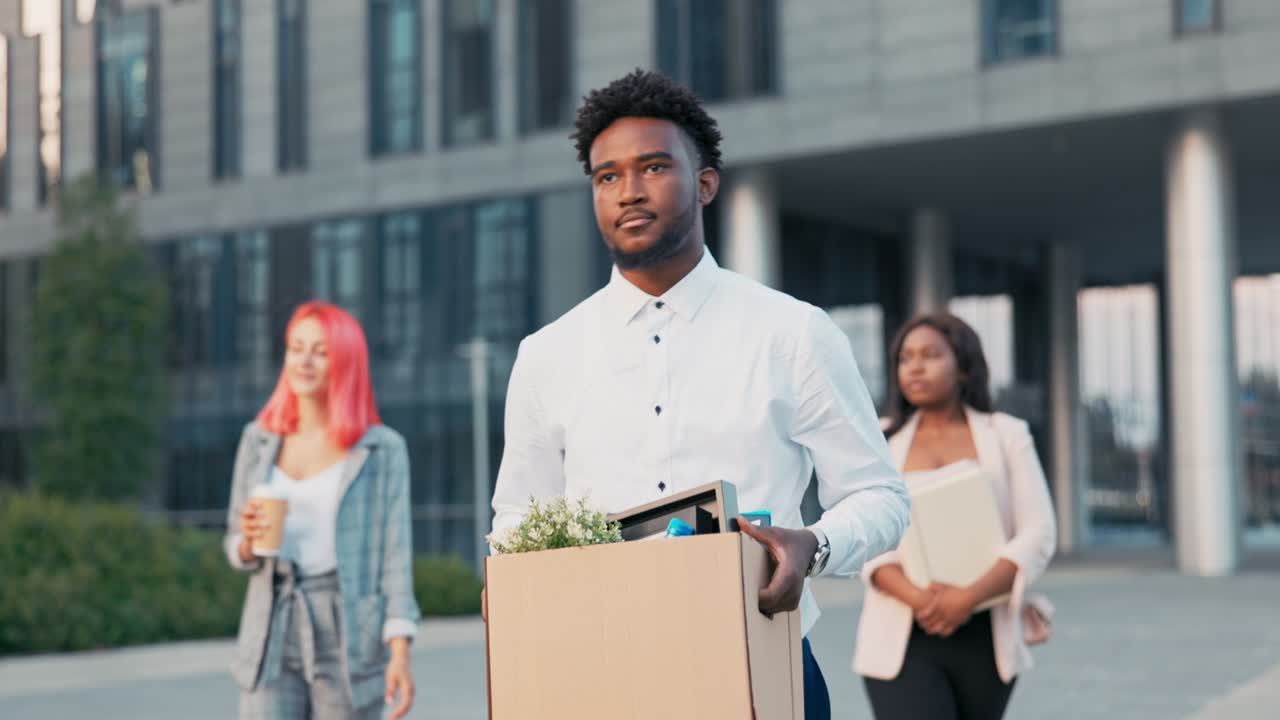 A dark-skinned man long-time office worker is fired from job, walks out of company building with co-workers, holding box of packed belongings in hands, a disgruntled expression, anger, sadness