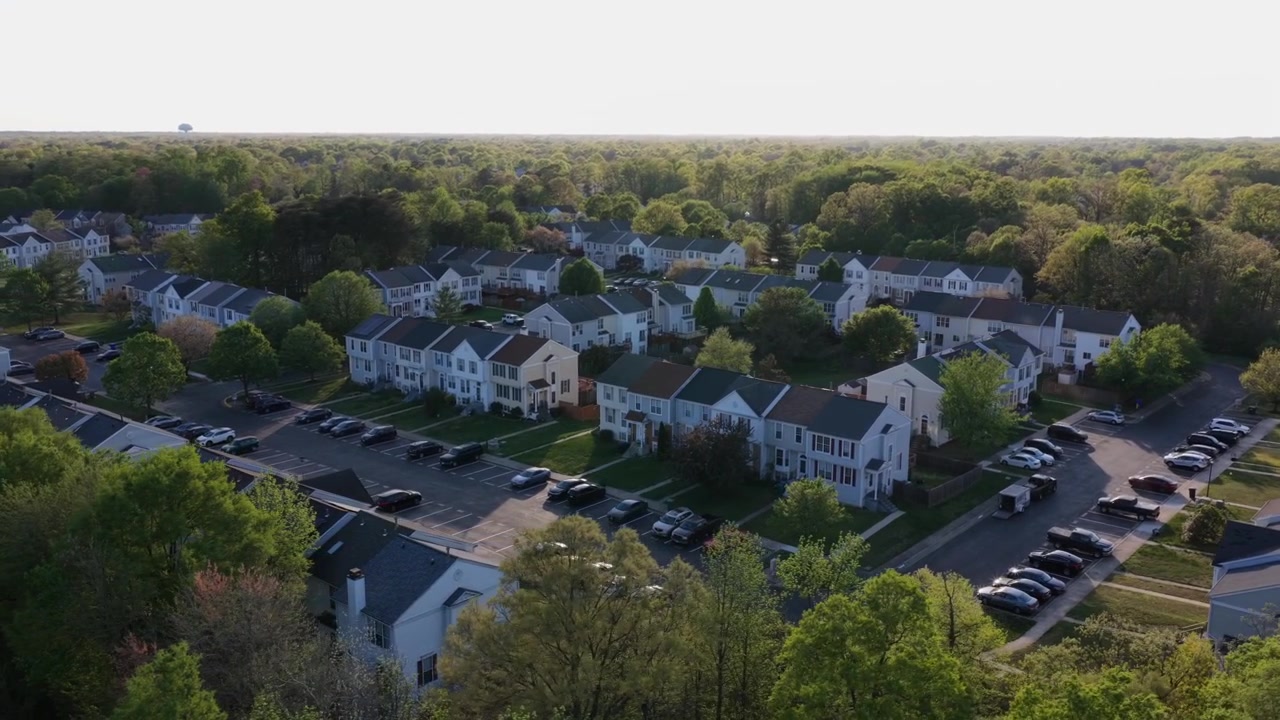Case a schiera suburbane a Waldorf, Maryland, al tramonto con alberi primaverili e tranquille strade suburbane. Residenze a due piani e auto parcheggiate si trovano in un paesaggio di quartiere tranquillo. Ampia ripresa aerea orbitale
