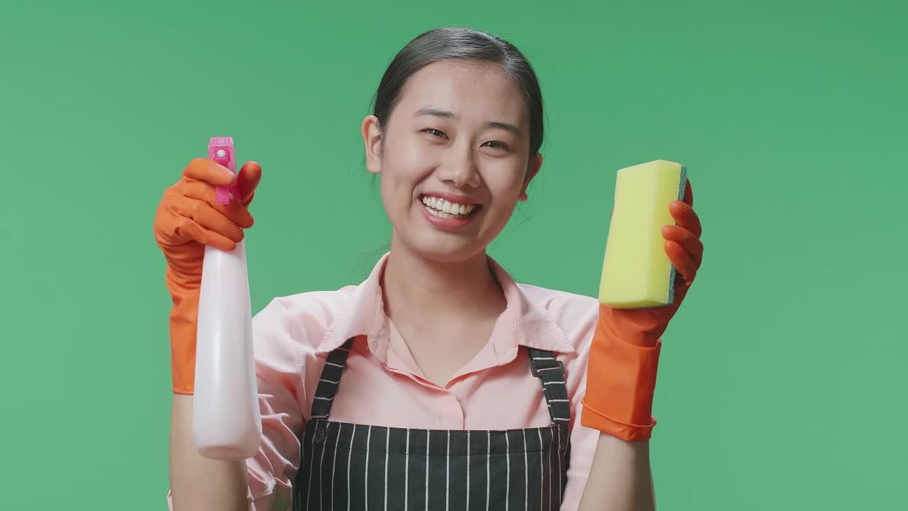 녹색 스크린 스튜디오 에 서 있는 스프레이 와 주방 스폰지 를 보여 주는 에프론 을 입은 미소 짓는 아시아 여성 집주인 의 근접 사진