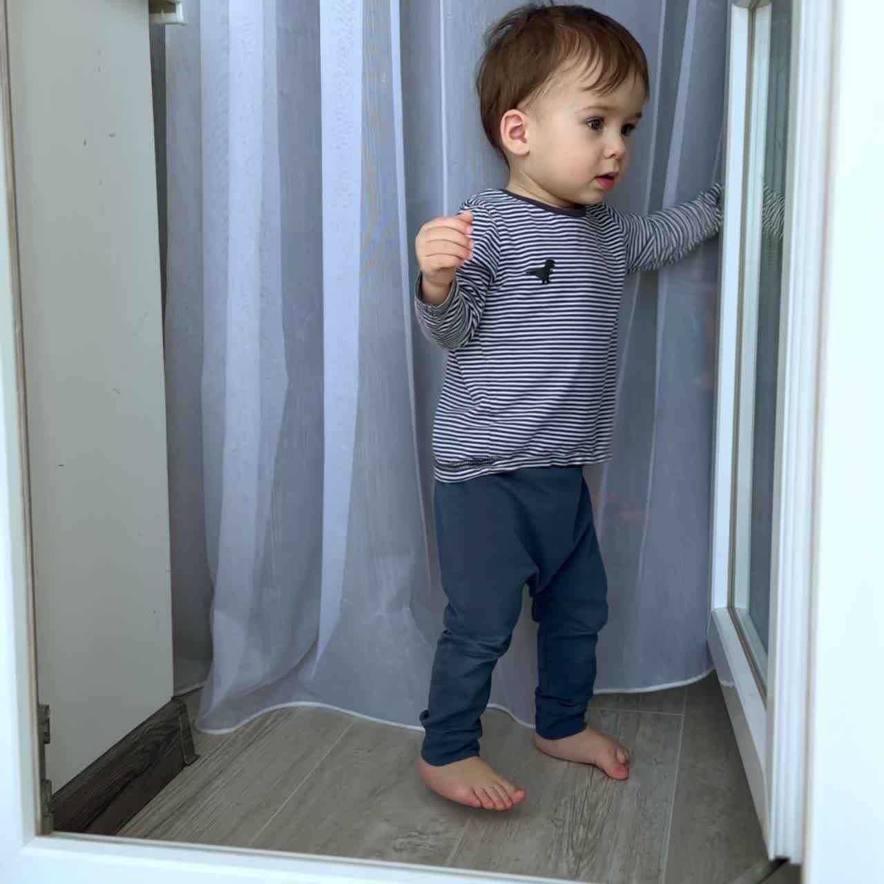 Sweet Caucasian baby boy stands at the balcony door and white curtain. Curious kid moves the door to and fro. Vertical screen
