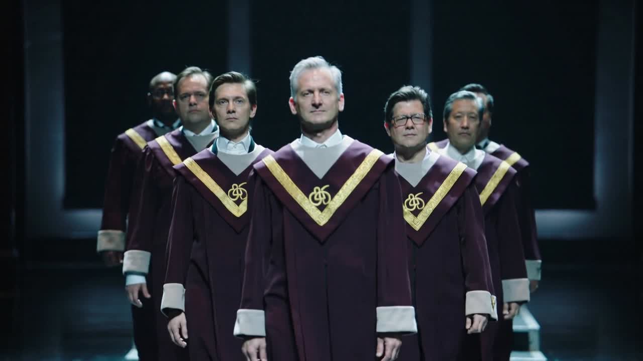 Marcherende mandskor bevæger sig fra platform, går ned ad trin på scenen, optræder i bordeauxfarvede kåber.