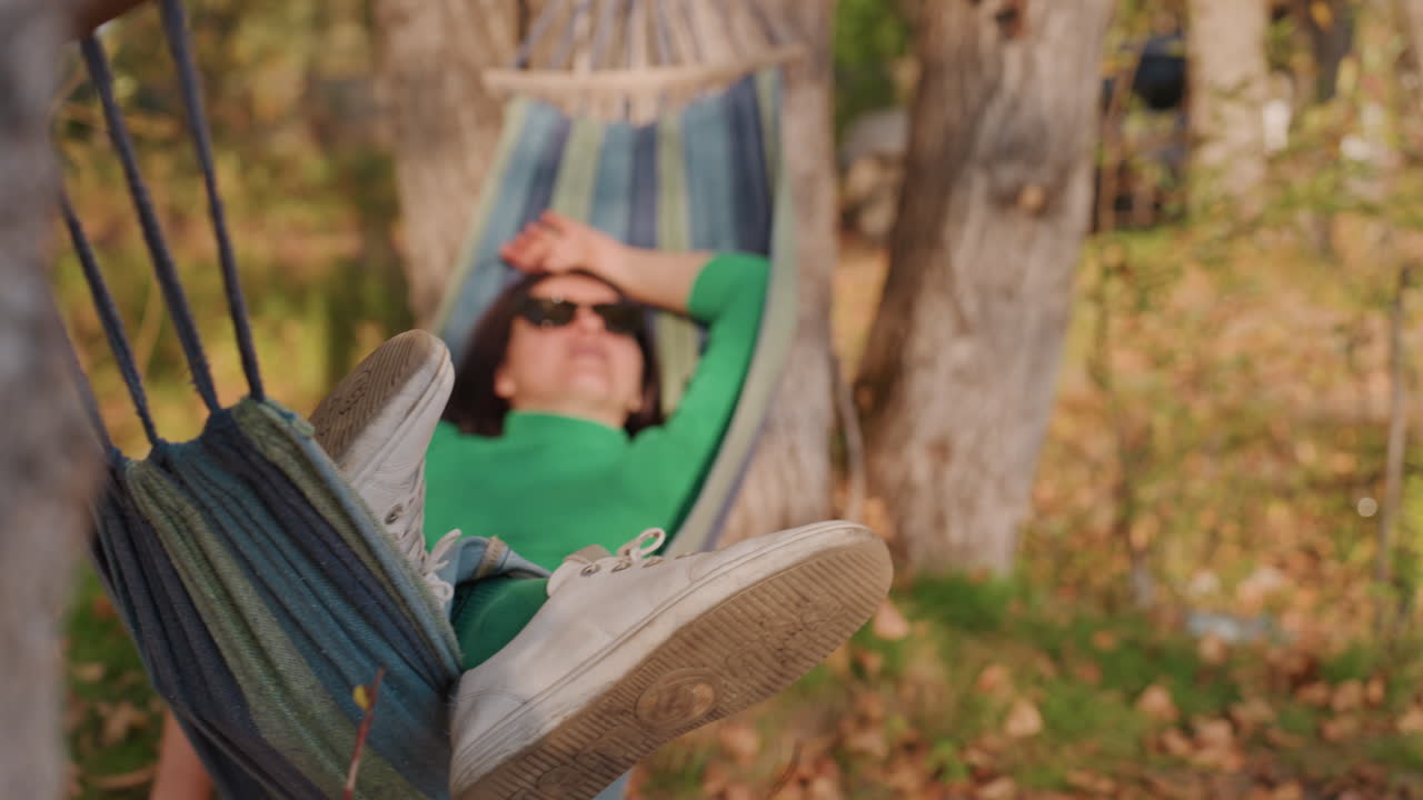 Persona in campeggio riposa in amaca tra gli alberi, con maglione verde e occhiali da sole. La dolce luce del sole filtra tra le foglie autunnali. L'amaca di corda è legata ai tronchi, un momento di quiete e riposo. Primo piano del busto e delle scarpe, con un lento e sereno dondolio.