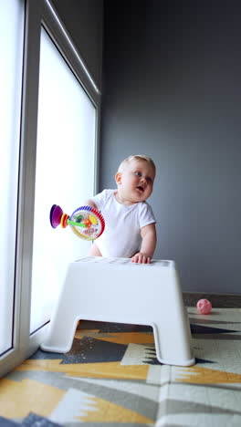 cute blond baby in white bodysuit stands near the chair playing with toy stuck to the window. happy kid laughs and pushes the chair. vertical video.