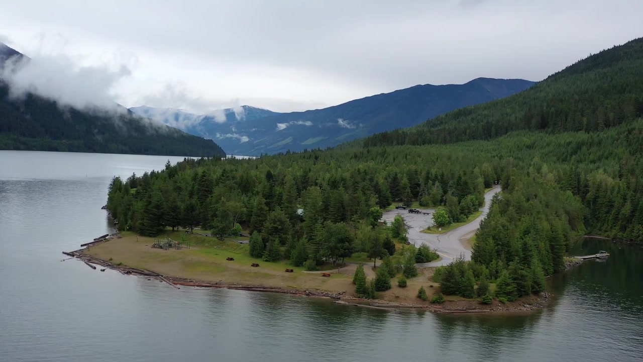 Ripresa cinematografica con drone dall'alto di una strada stretta che si snoda attraverso una fitta foresta di conifere accanto a un lago calmo e riflettente. Cime montuose suggestive e nuvole basse definiscono il paesaggio della Columbia Britannica.