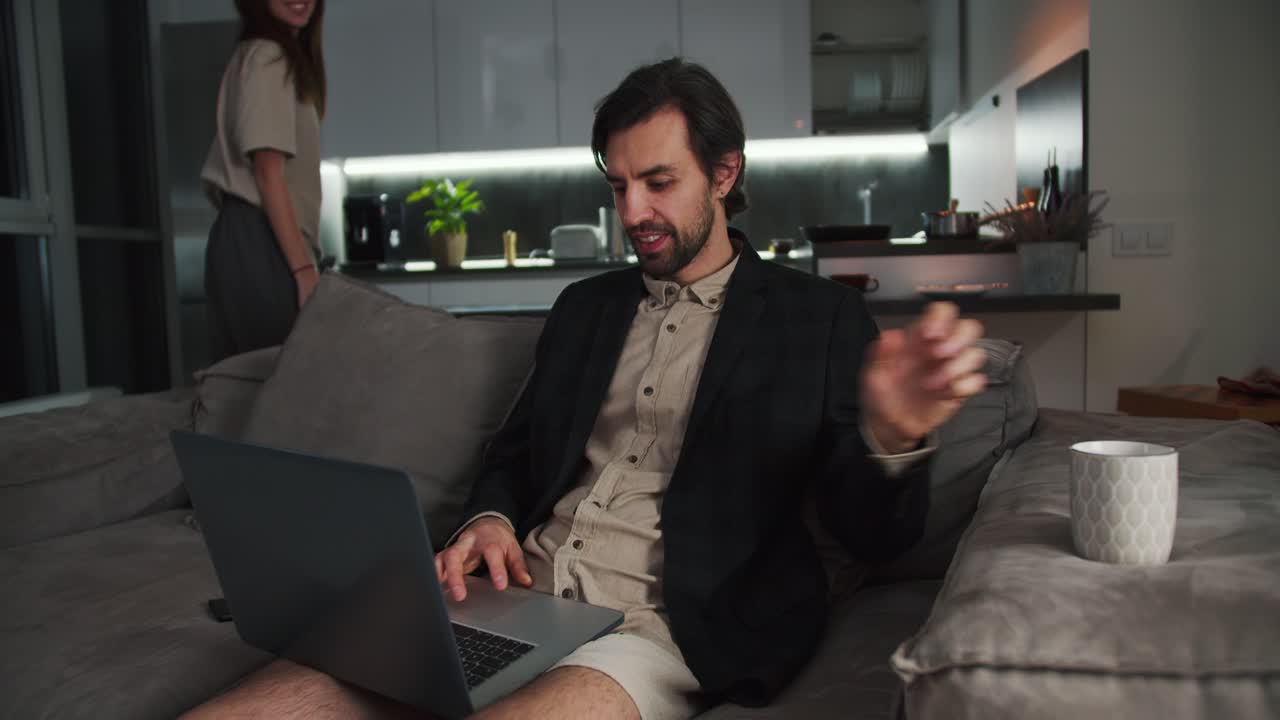A brunette man with stubble in a black jacket and beige shorts sits on the sofa and works at a gray laptop drinking tea while his brunette girlfriend hugs his shoulders from behind in a modern studio apartment in the evening