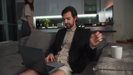 un hombre moreno con paja en una chaqueta negra y pantalones cortos beige se sienta en el sofá y trabaja en una computadora portátil gris bebiendo té mientras su novia morena abraza sus hombros por detrás en un apartamento estudio moderno por la noche