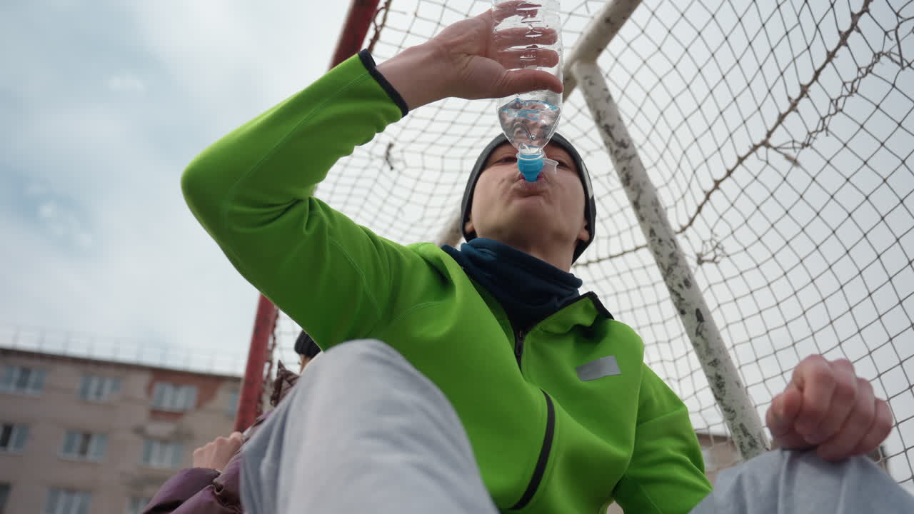 Hombre caucásico con camiseta verde bebiendo agua de una botella en ángulo bajo cerca de la portería, perspectiva del portero con la red y edificios urbanos al fondo, pausa intensa de un atleta durante el entrenamiento