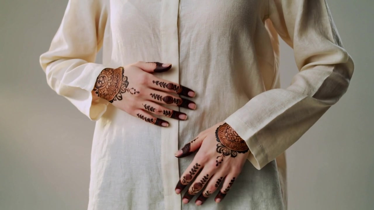 Iniziando con le mani sul petto, una donna con una tunica color crema sposta le mani su una parete semplice, mettendo in mostra l'henné.