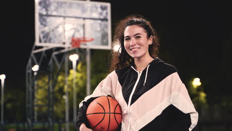 retrato de una hermosa jugadora de baloncesto sosteniendo una pelota y sonriendo a la cámara en una cancha al aire libre por la noche