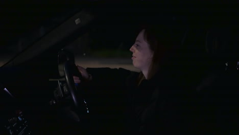 Happy-woman-girl-listening-to-music-in-car-parked-at-side-of-busy-road-at-night
