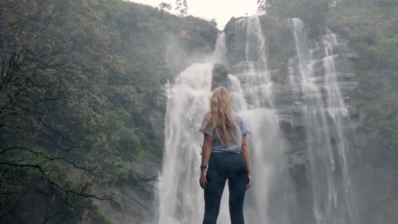 A woman stands in awe before the roaring Bomburu Ella Waterfalls, surrounded by mist, jungle, and rocky cliffs in the lush highlands of Nuwara Eliya, Sri Lanka.