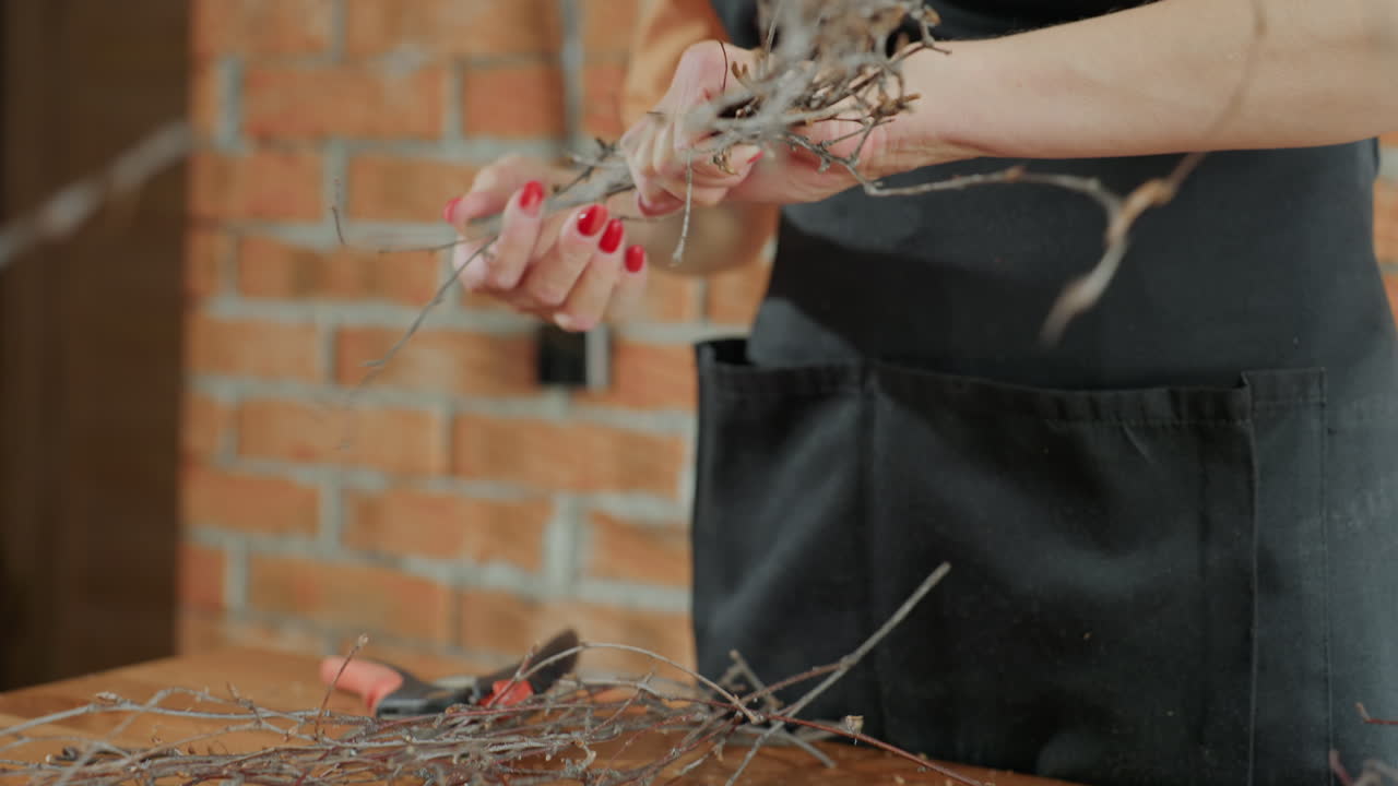Female florist hands cutting dry twigs with pruning shears on wooden table during floral arrangement preparation, creating rustic decoration with natural materials inside creative studio