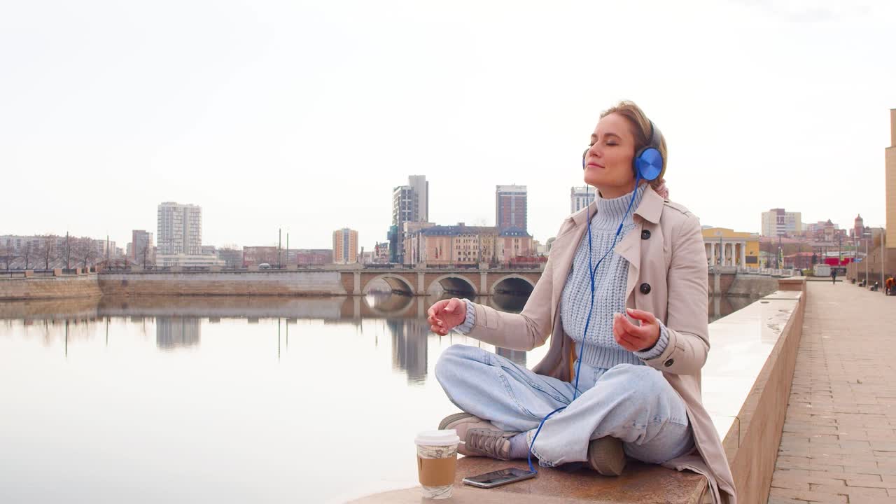 A young woman meditates on the embankment. She sits in headphones and listens to relaxing music. Rest from office bustle and city noise