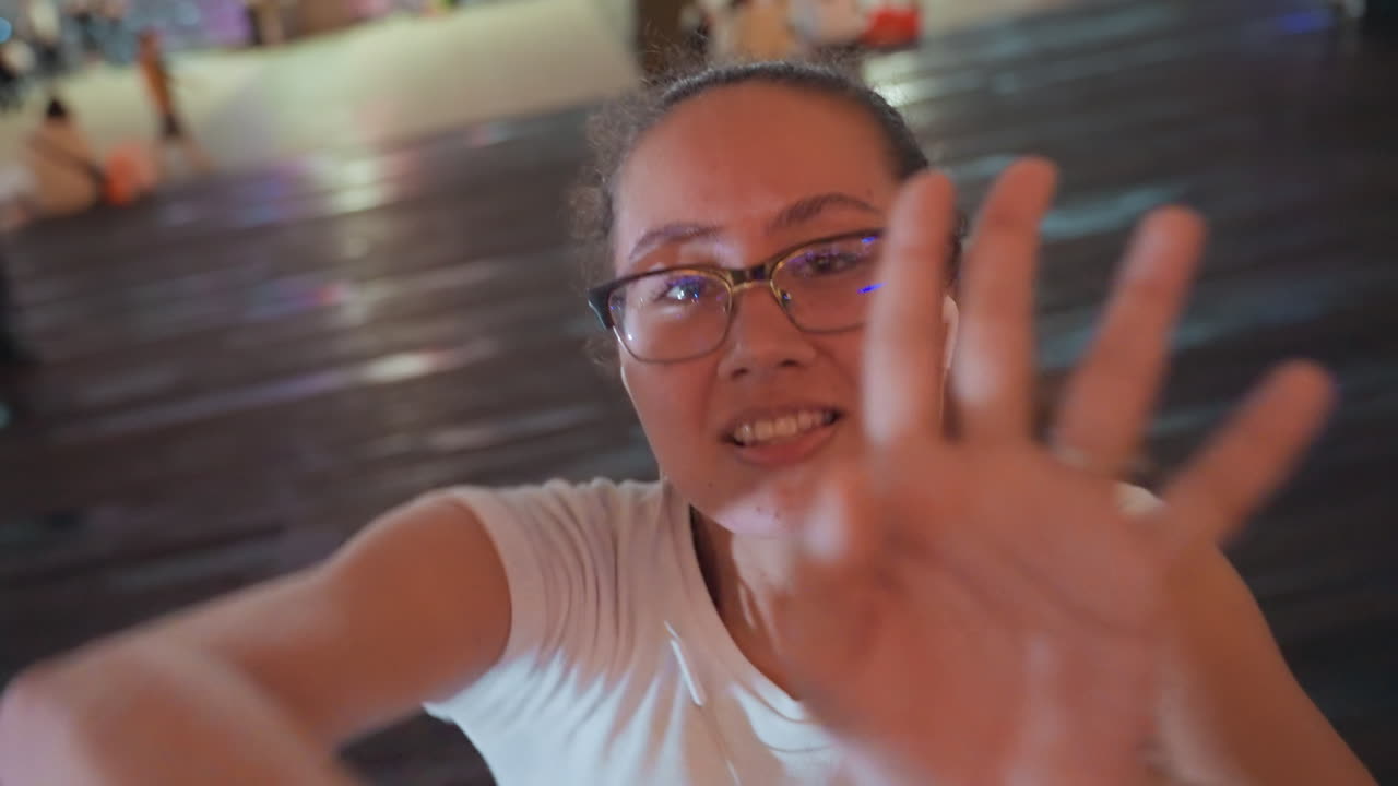 Vlogger donna giocosa che si protende verso la fotocamera su una passerella, che balla e gesticola in una città illuminata al neon, con occhiali e auricolari, maglietta casual, espressione allegra, folla serale e luci sfocate, riprese selfie spontanee