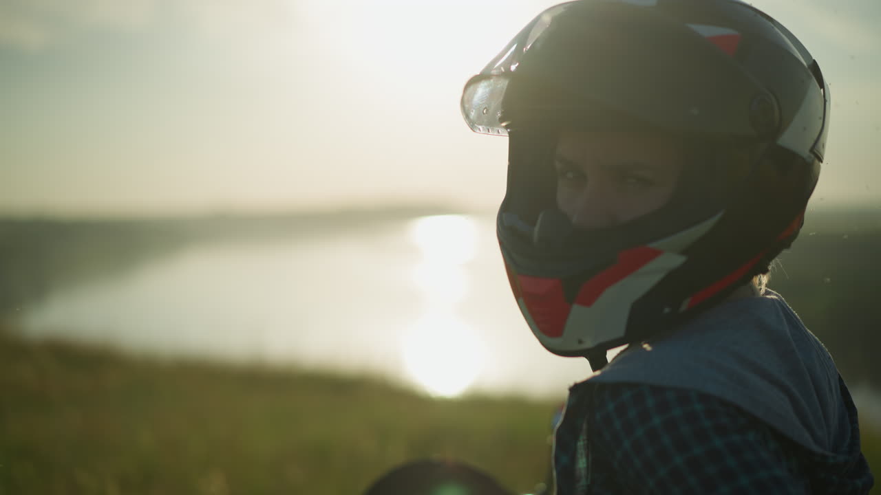 A woman in a helmet sits on a motorcycle in a grassy field, looking out towards the camera, with a blur view of water body in the distance
