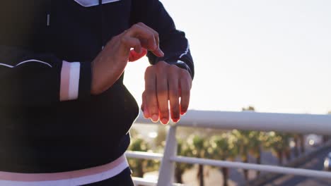 Midsection-of-african-american-man-exercising-on-footbridge,-checking-smartwatch,-backlit-by-sun