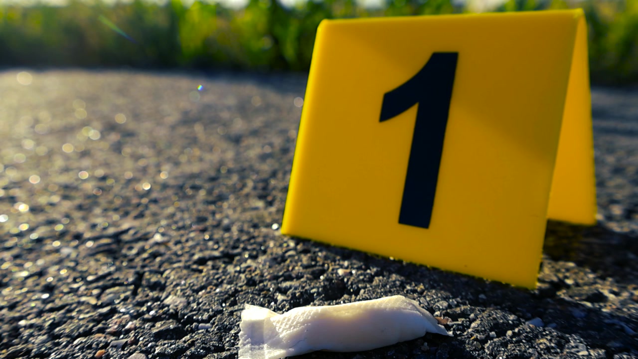 Primo piano di un marcatore giallo per prove forensi con il numero uno, posizionato accanto a una piccola bustina bianca di nicotina su una strada asfaltata durante un'indagine di polizia, con uno sfondo sfocato