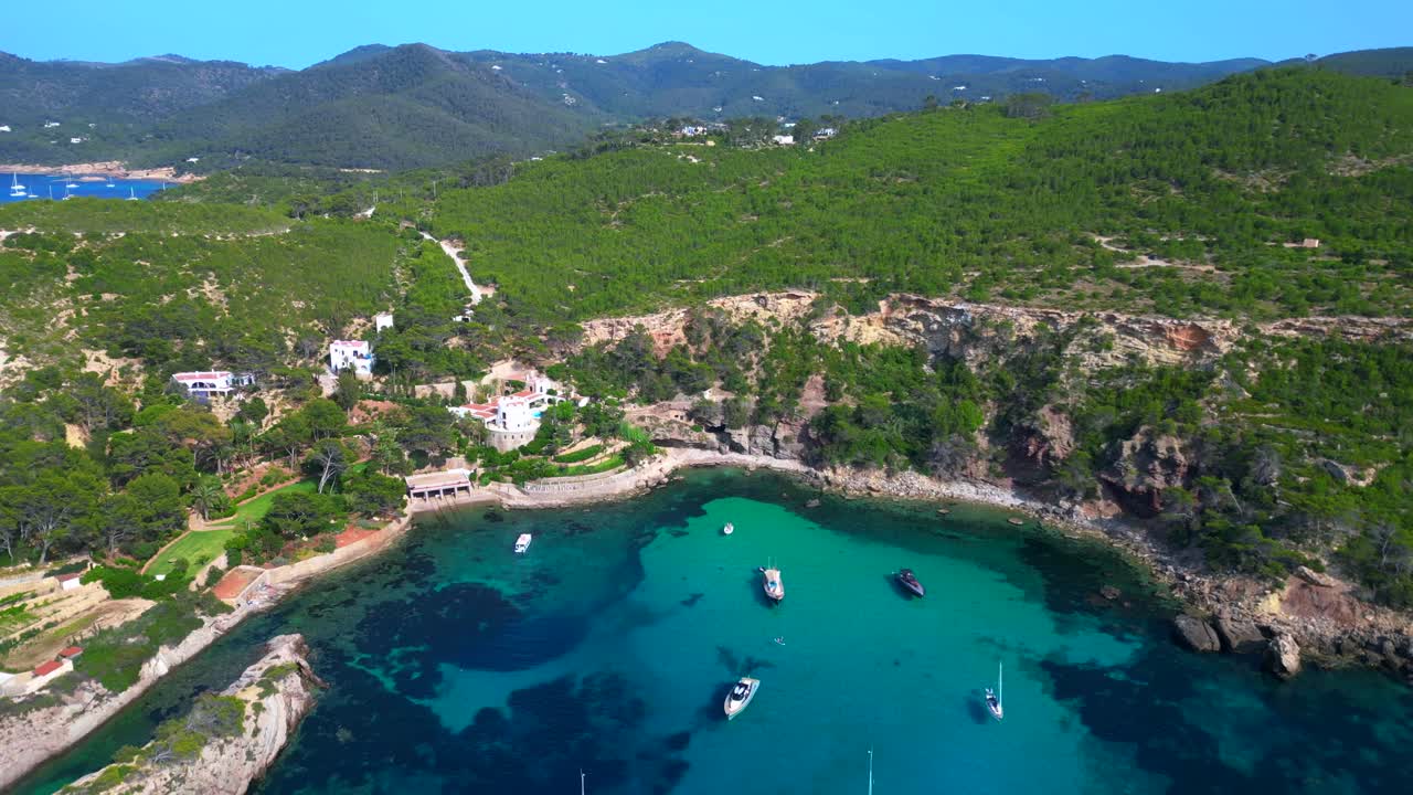 luxury villa in Ibiza, Spain, overlooking a picturesque bay with turquoise water and boats sailing on a sunny summer day. Nice aerial view flight fly reverse drone