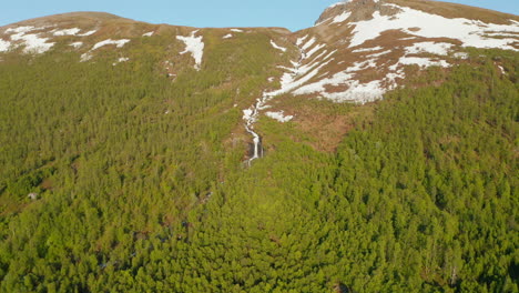 Aerial-view-towards-a-waterfall-on-a-snowy-mountain,-sunny,-summer-day,-in-Rotsund,-Troms,-Nordland,-Norway---descending,-dolly,-drone-shot