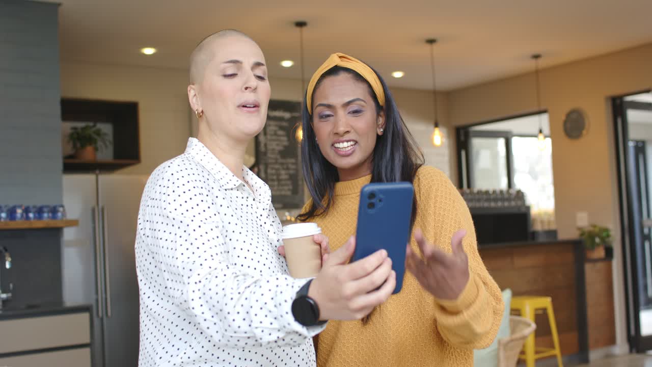 Diverse female friends holding blue smartphone, recording lip-sync while gesturing cup in cafe