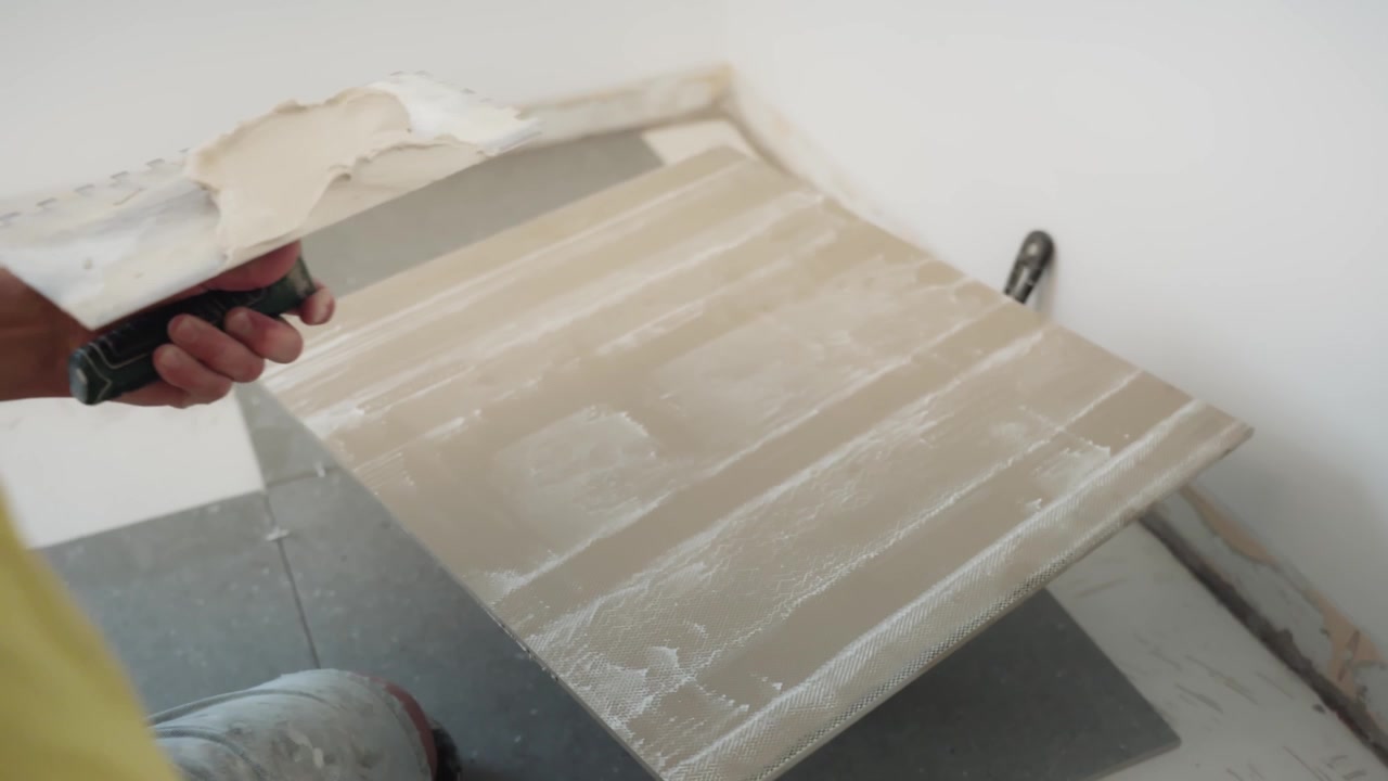 Close-up of a worker scooping and spreading fresh adhesive onto the back of a large floor tile with a trowel during interior installation.
