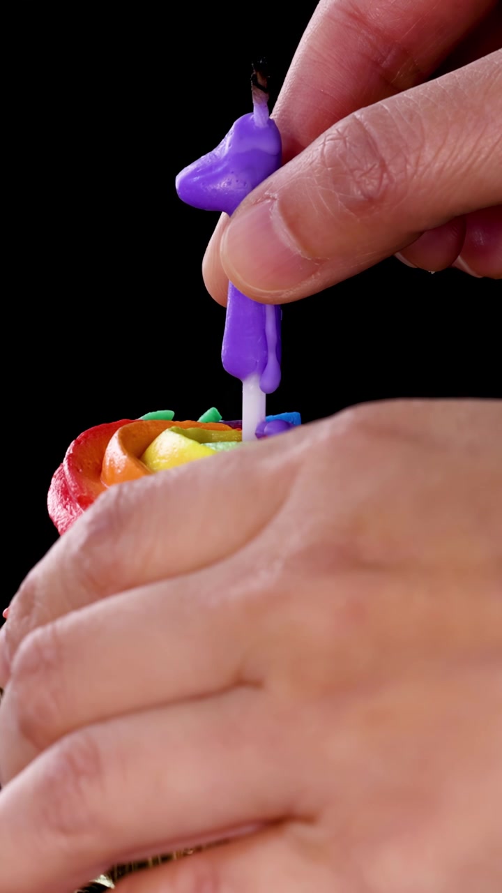 Uma mão insere uma vela roxa com o número um em um cupcake com cobertura colorida de arco-íris contra um fundo preto, com enquadramento de câmera em close-up, bem iluminado e estável.