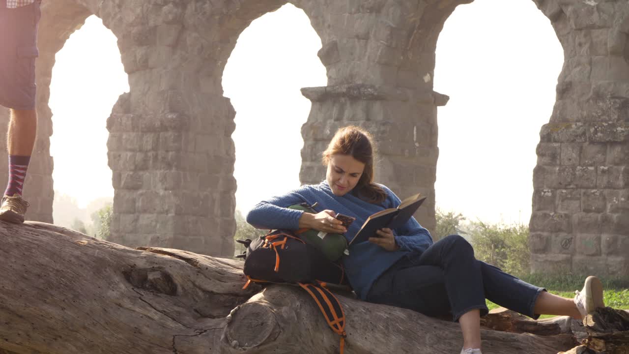 pareja feliz de mochileros turistas en un tronco de madera tocando la guitarra cantando leyendo un libro frente al antiguo acueducto romano ruinas en el romántico parque degli aquedotti parque en roma al amanecer cámara lenta
