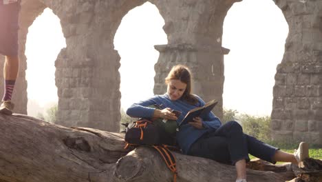 happy young couple backpackers tourists on a log trunk playing guitar singing reading book in front of ancient roman aqueduct ruins in romantic parco degli acquedotti park in rome at sunrise slow motion