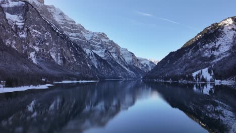 stunning aerial drone pulls back klöntalersee in winter with pristine reflections of snowy swiss alps surrounded by serene natural beauty.