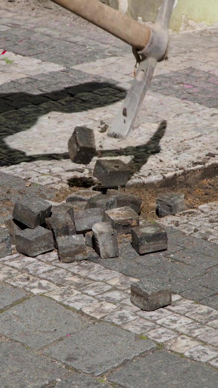 A worker uses a pickaxe to pry up stone pavers from a sunny sidewalk.