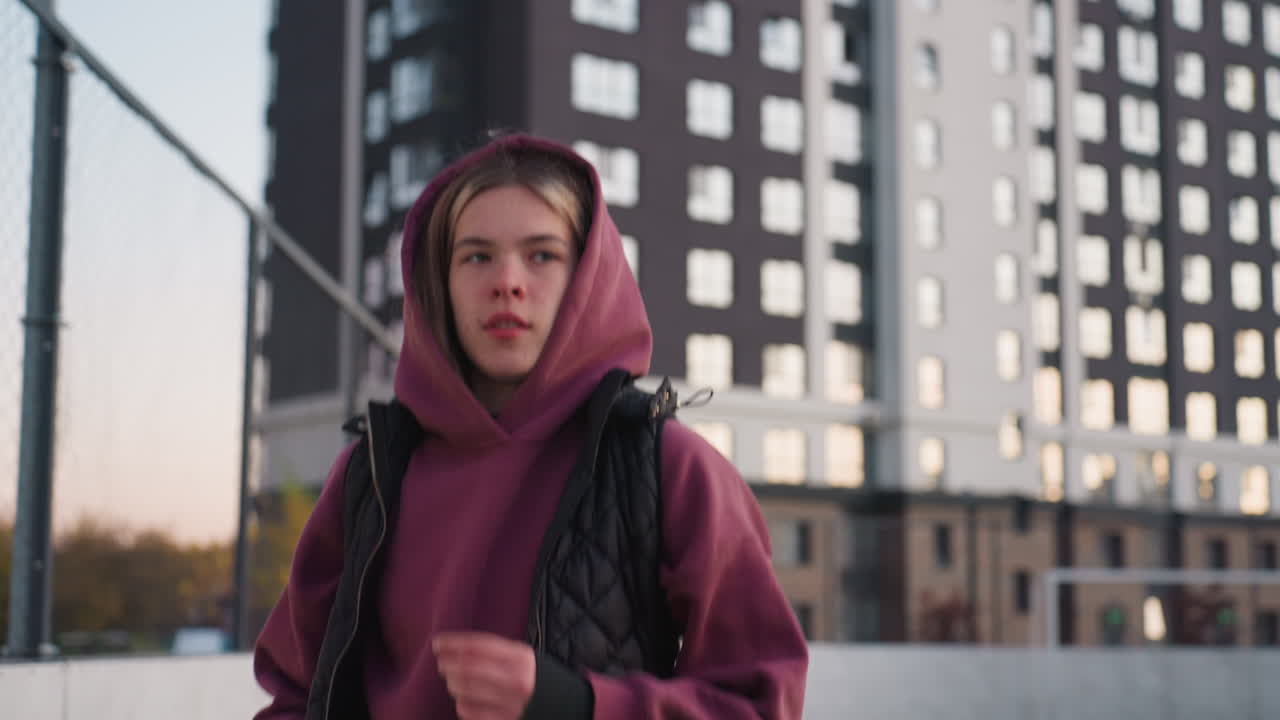 Energetic female jogger in hoodie runs around curved outdoor barbed wire sports court under clear dusk sky, framed by chain link fence with backdrop of apartment blocks lit by soft sunset glow