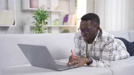 Un-Joven-Africano-Reflexivo-Que-Trabaja-Desde-Casa-Mirando-Una-Computadora-Portátil-Y-Tomando-Notas-En-Papel.