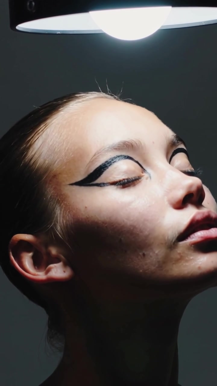 Vertikal video: Model, der ruller hovedet og vipper hagen i minimalistisk top under lampe, viser sort eyeliner