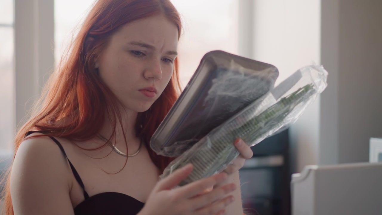 Donna bianca dai capelli rossi legge una lettera alla scrivania, fronte corrugata ed espressione concentrata, busta e documenti sparsi vicino alla finestra, sfondo di appartamento urbano, umore preoccupato, scena di revisione documenti