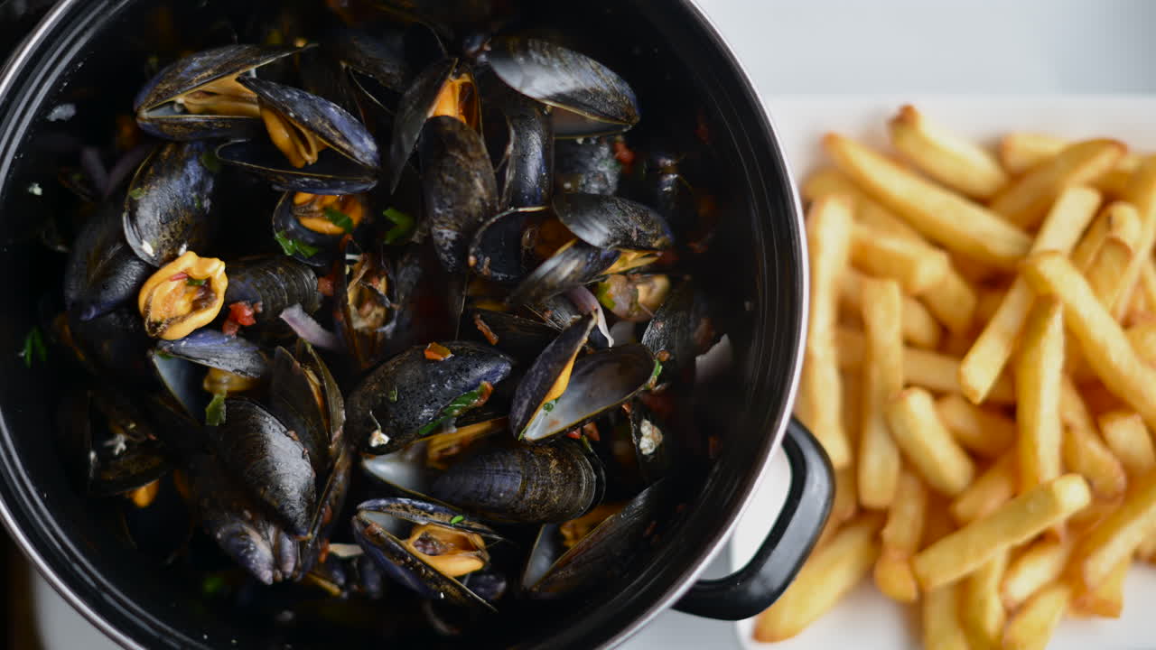 Cooked mussels served in a pot with herbs and sauce, accompanied by golden fries on the side