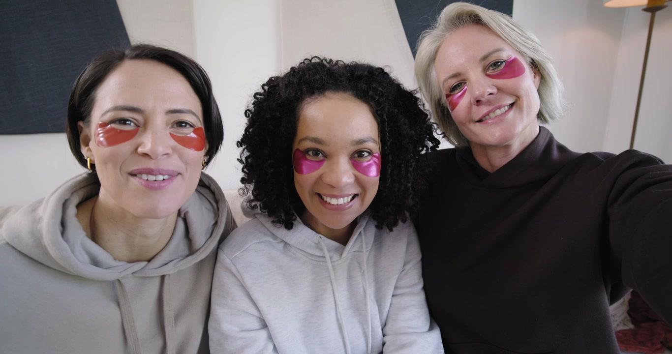 Amiche di diverse etnie che si salutano sul divano mentre la donna a destra inizia un selfie, toccandosi i patch sotto gli occhi.