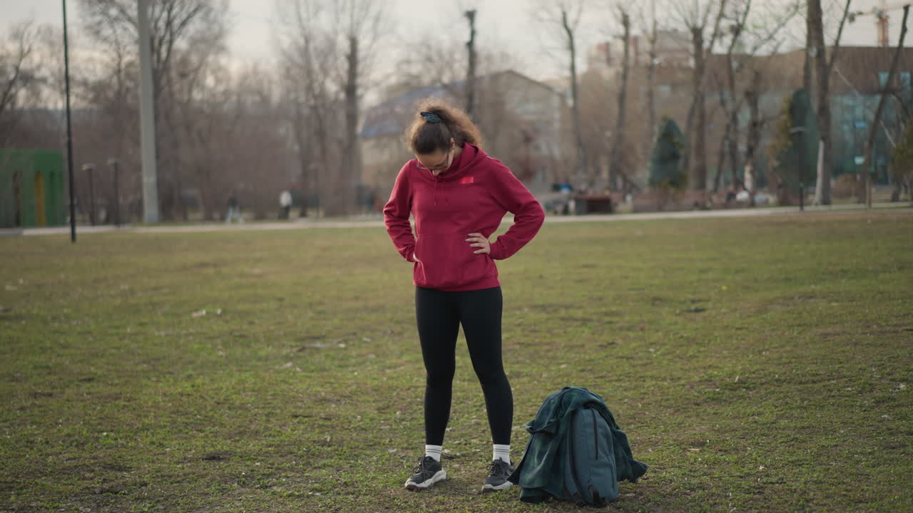 Youthful Woman Assessing Posture, Female In Red Hoodie Examining Her Stance Outdoors, Teenage Female Wearing Vivid Hoodie Checks Her Posture Attentively Beside Backpack In Lush Park Environment