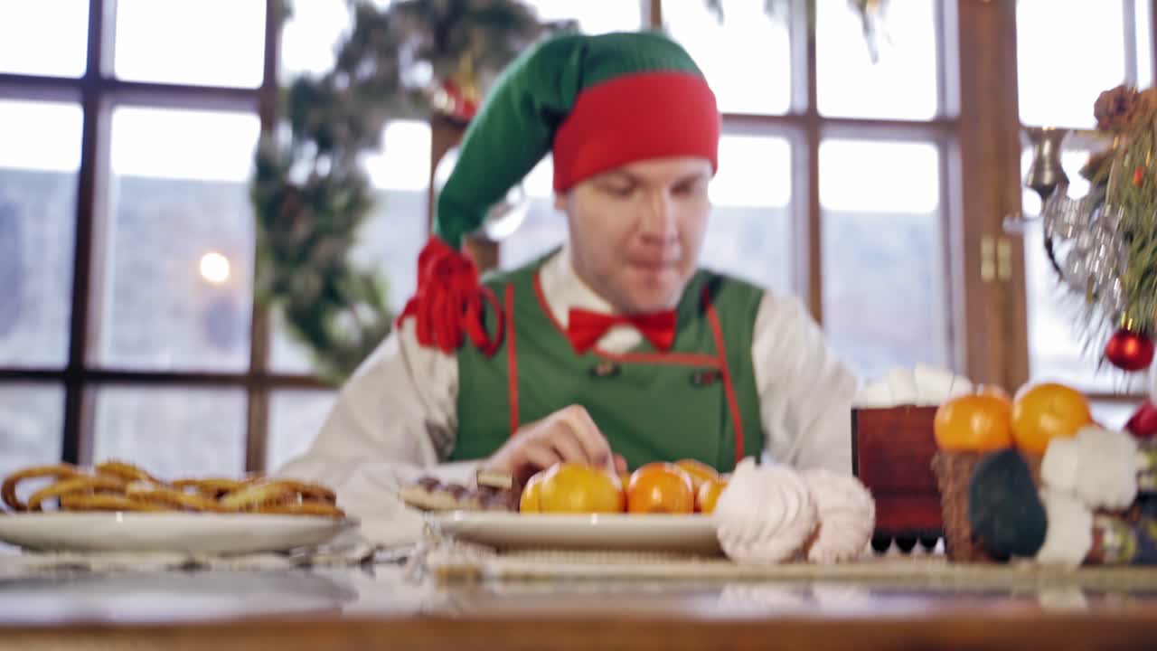 Hungry elf is impatiently tapping his feet at the Christmas table. Male elf in green costume with hood is sitting at festive table in front of the oranges. New Year and Christmas concept.