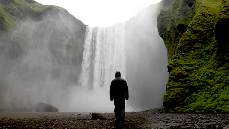 Cascadas-De-Skogafoss-En-Islandia-Con-Un-Hombre-Con-Chaqueta-De-Lluvia-Caminando-Hacia-Las-Cataratas-En-Cámara-Lenta