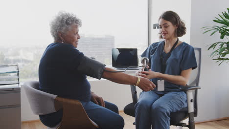 Nurse-Wearing-Scrubs-In-Hospital-Office-Checking-Senior-Female-Patients-Blood-Pressure