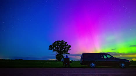 timelapse de coloridas luces del norte con árbol y furgoneta en primer plano con el hombre sentado en una silla